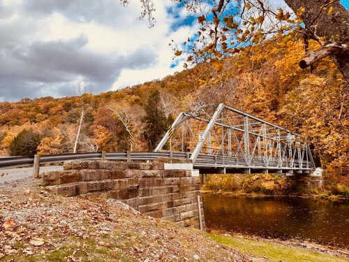 A metal bridge spans a river, surrounded by autumn foliage and a cloudy sky. Leaves cover the ground nearby.