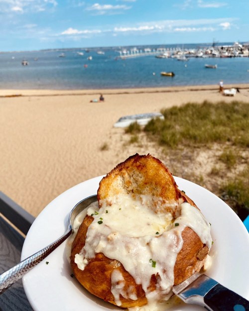 A bowl of creamy soup in a bread bowl, with a beach and boats in the background under a blue sky.