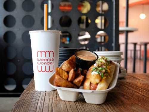 A tray with a hot dog topped with toppings, potato wedges, a cup of sauce, and a drink from Weenie Wonder.