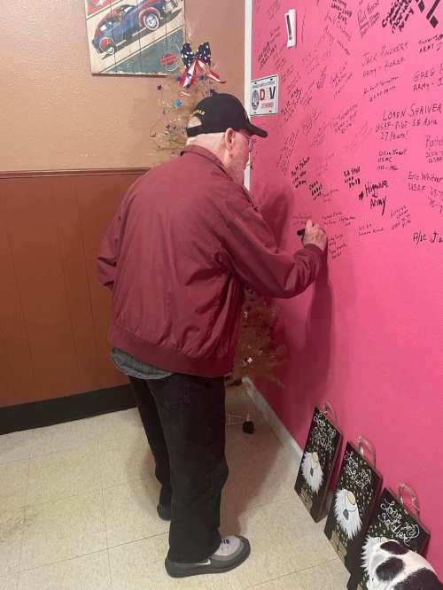An older man in a maroon jacket writes on a pink wall decorated with signatures, next to a small Christmas tree.