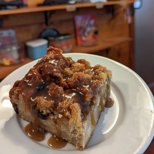 A slice of dessert with a crumbly topping and caramel drizzle, served on a white plate against a rustic background.
