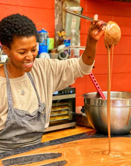 A woman with short hair smiles as she stirs melted chocolate in a kitchen, with a bowl and utensils nearby.