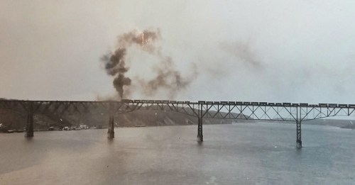 A vintage photo of a bridge over water, with smoke rising from a train crossing it.
