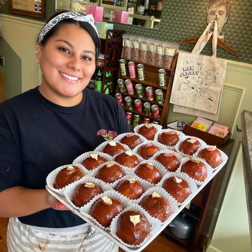 A smiling woman holds a tray of chocolate-covered treats in a cozy shop filled with snacks and decor.