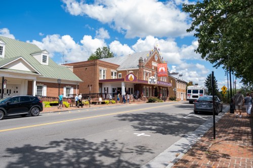 A street view of a town with a theater, people walking, and cars parked along the road under a blue sky with clouds.