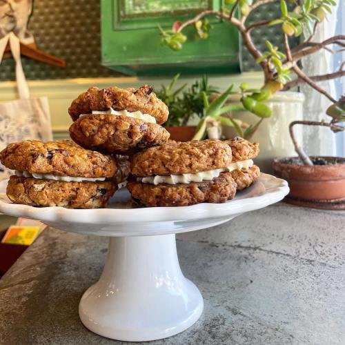 A plate of oatmeal cookies with cream filling, displayed on a white pedestal, surrounded by plants.