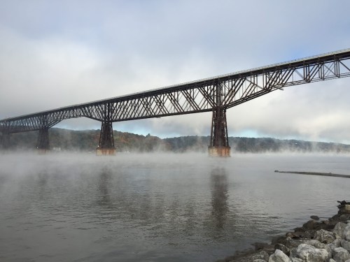 A large bridge spans a foggy river, with rocky banks and mist rising from the water beneath a cloudy sky.