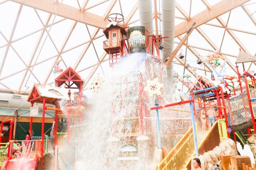 A colorful indoor water park with a large water slide and a waterfall feature, surrounded by play structures.