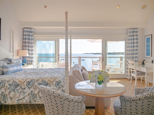 Bright hotel room with a sea view, featuring a bed, seating area, and a table with flowers. Large windows let in natural light.