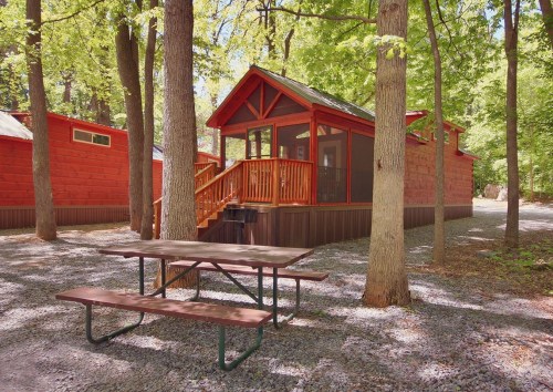 A red cabin with a screened porch surrounded by trees, with a picnic table in the foreground on a gravel path.