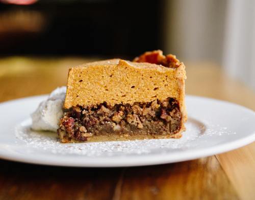 A slice of pumpkin pie layered with a pecan crust, served on a white plate with a dollop of whipped cream.