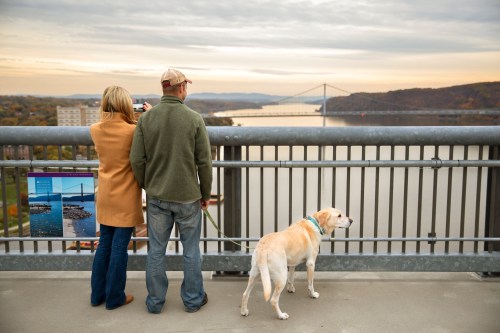 A couple and their dog enjoy a scenic view of a river and bridge at sunset.