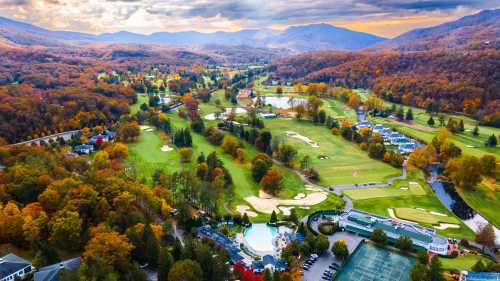 Aerial view of a scenic golf course surrounded by autumn foliage and mountains under a cloudy sky.