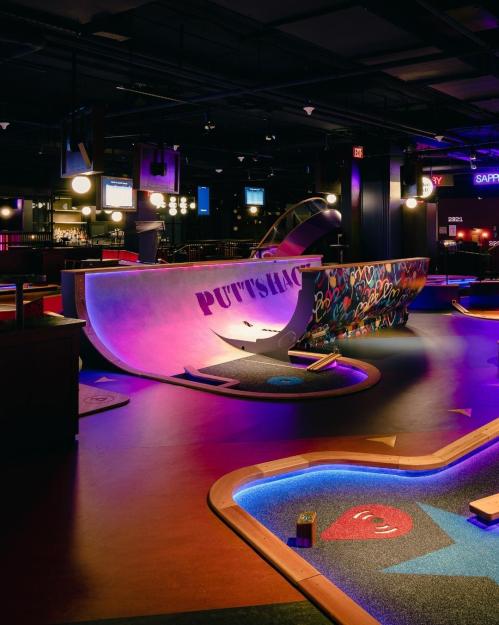 A vibrant indoor mini-golf course with colorful lighting and unique obstacles, featuring a graffiti wall and bar area.