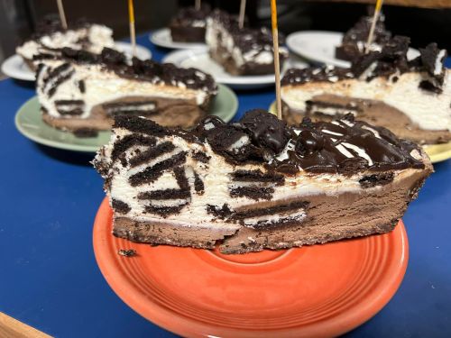 A slice of layered chocolate and cream cake topped with crushed cookies, served on a red plate.