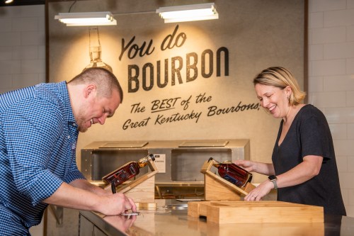Two people tasting bourbon at a bar, with a sign in the background that reads "You do Bourbon."
