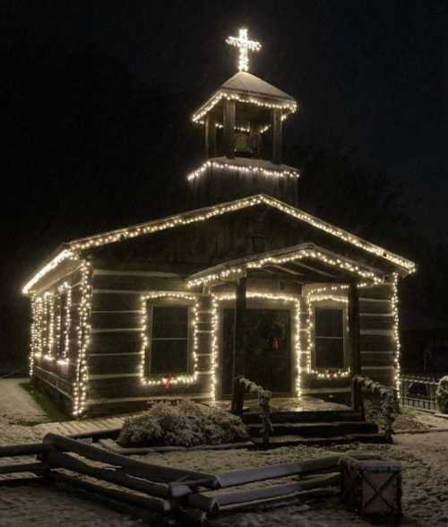 A log cabin church illuminated with festive lights, featuring a cross on top, surrounded by snow at night.