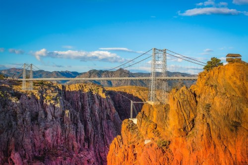 A suspension bridge spans a deep canyon, surrounded by rocky cliffs and distant mountains under a blue sky.