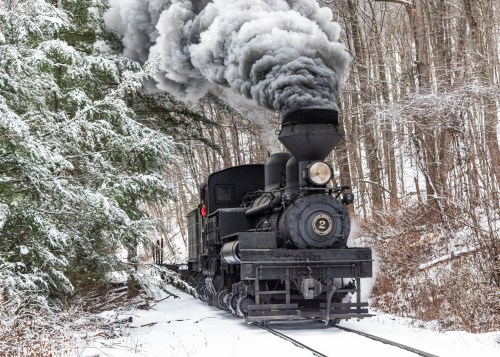 A vintage steam train emits dark smoke while traveling through a snowy forest landscape.