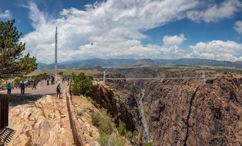 A scenic view of a canyon with a bridge, surrounded by mountains and a cloudy sky, with visitors exploring the area.