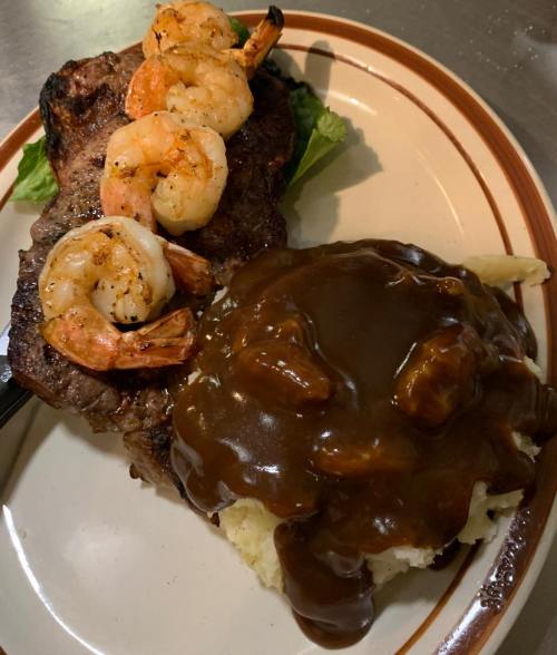 A plate featuring grilled shrimp atop a steak, served with mashed potatoes and brown gravy.
