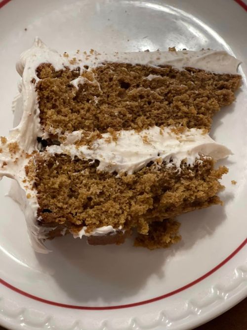A slice of cake with a light frosting on a white plate, showing a moist, textured interior.