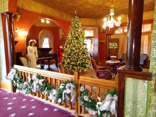 A festive room decorated for Christmas, featuring a large tree, a piano, and elegant furnishings.