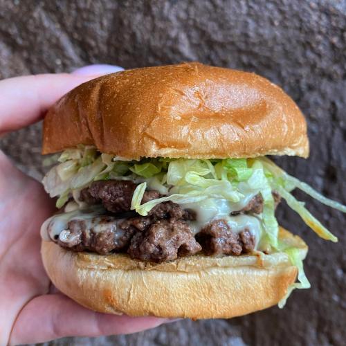 A hand holding a juicy burger with lettuce and melted cheese on a soft bun against a textured background.