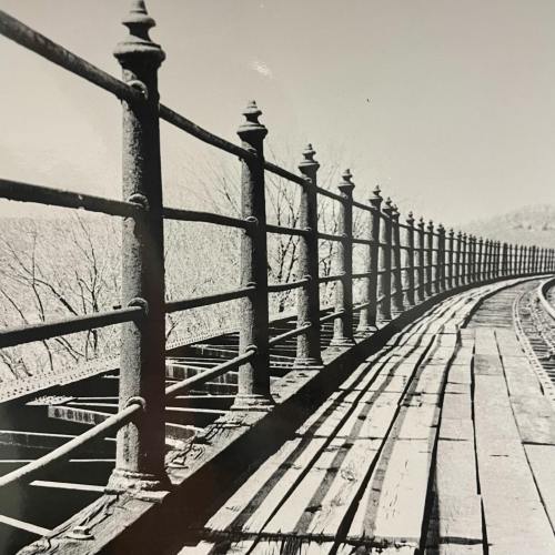 Black and white image of a wooden walkway with a metal railing, leading into the distance.