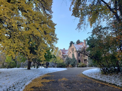 A scenic view of a path leading to a large house, surrounded by autumn trees and patches of snow.