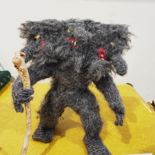 A fuzzy, three-headed creature with red eyes holds a stick, standing on a yellow surface.