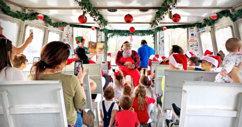A festive scene on a boat with children and adults in Santa hats, gathered around Santa reading a story.