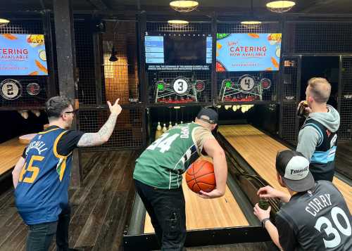 Four friends in sports jerseys are bowling while one prepares to roll a basketball down the lane. Screens are visible above.