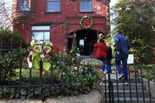 People enter a festive brick building decorated for the holidays, with wreaths and a large "S" on the door.