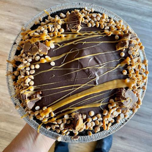 A hand holding a round chocolate pie topped with caramel drizzle, peanut butter cups, and crunchy toppings.