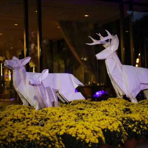 Two white deer sculptures among vibrant yellow flowers, set against a softly lit indoor background.