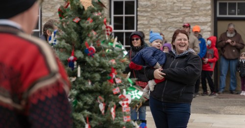 A woman smiles while holding a baby, with a decorated Christmas tree and a crowd in the background.