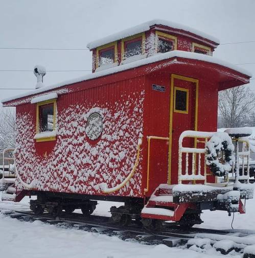 A bright red caboose covered in snow, with a wreath on the front, set against a winter landscape.