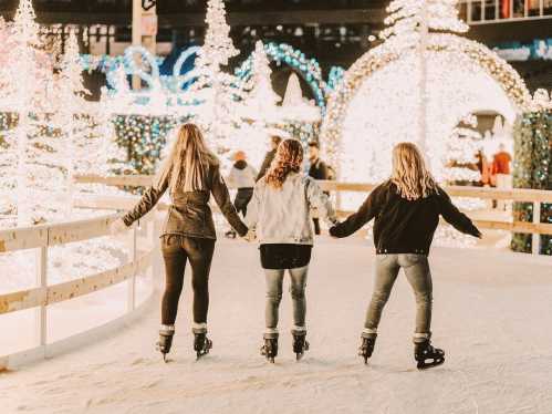 Three girls ice skating hand in hand, surrounded by festive lights and decorations.