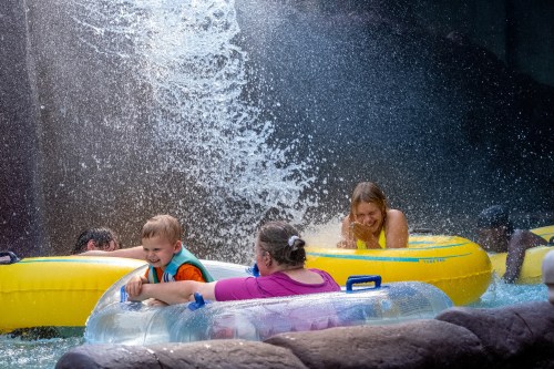 A group of people, including a child, enjoy a water ride with splashes and yellow inner tubes in a fun, sunny setting.