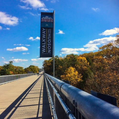 A walkway over a river with a banner reading "Walkway Over the Hudson" and trees in the background under a blue sky.