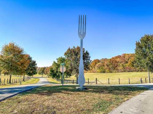 A large fork sculpture stands at the end of a pathway, surrounded by trees and a clear blue sky.