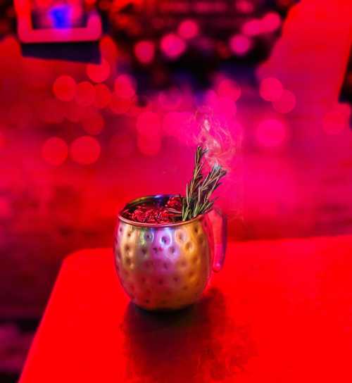 A metallic mug filled with a cocktail, garnished with rosemary, surrounded by a vibrant pink background.