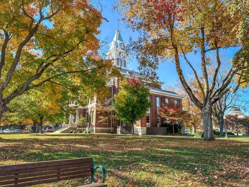 A historic building surrounded by colorful autumn trees and a clear blue sky, with a park bench in the foreground.