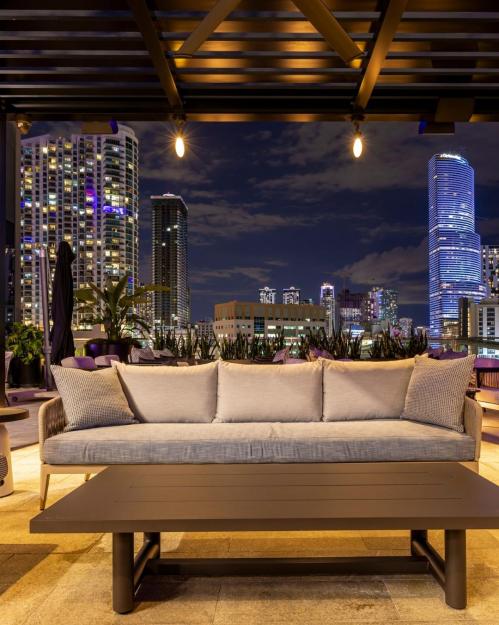 A modern outdoor lounge area with a sofa and table, overlooking a city skyline at night with illuminated buildings.