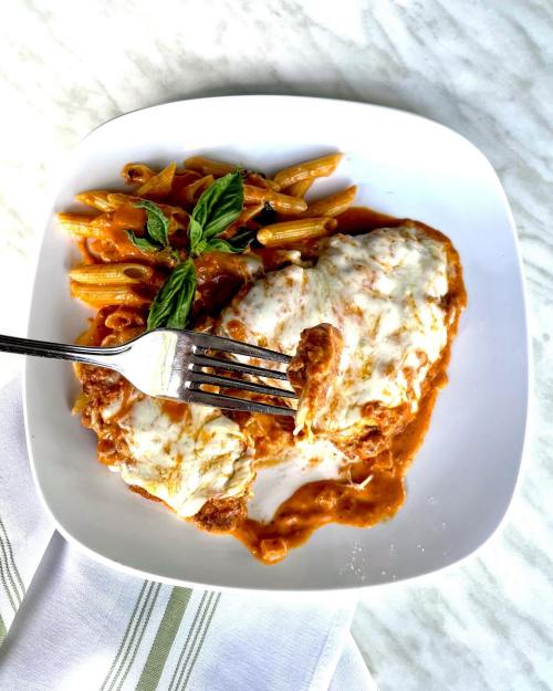 A plate of chicken parmesan with pasta, topped with melted cheese and fresh basil, served with a fork.