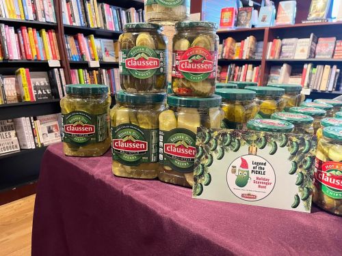 A display of Claussen pickle jars on a table in a bookstore, with a sign about a pickle-themed event.