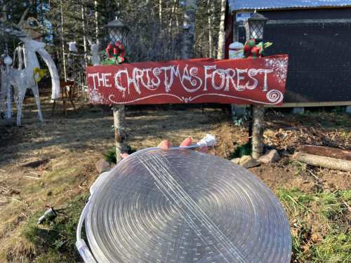 A sign reading "The Christmas Forest" with festive decorations, in a wooded area, with a roll of lights in the foreground.