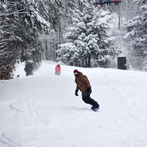 A snowboarder glides down a snowy slope, surrounded by trees, with another person in the background.