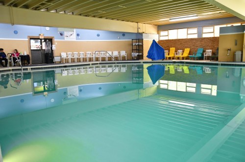 Indoor swimming pool with clear water, surrounded by chairs and a blue tarp, under bright lighting.